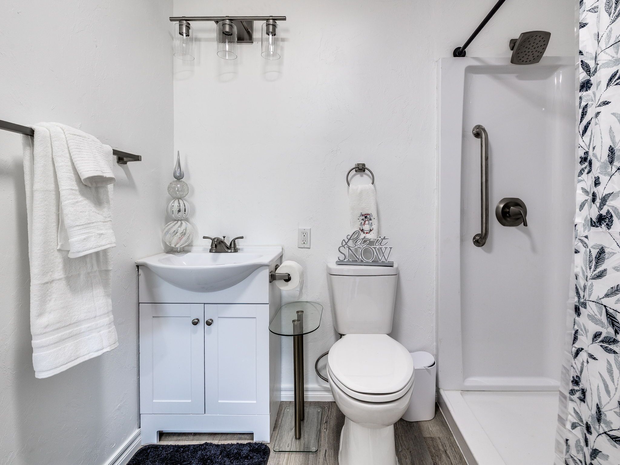 Un bany modern amb un lavabo, vàter i dutxa de color blanc. Hi ha un para-tovallons amb una tovallola penjada, i una porta de dutxa de vidre.