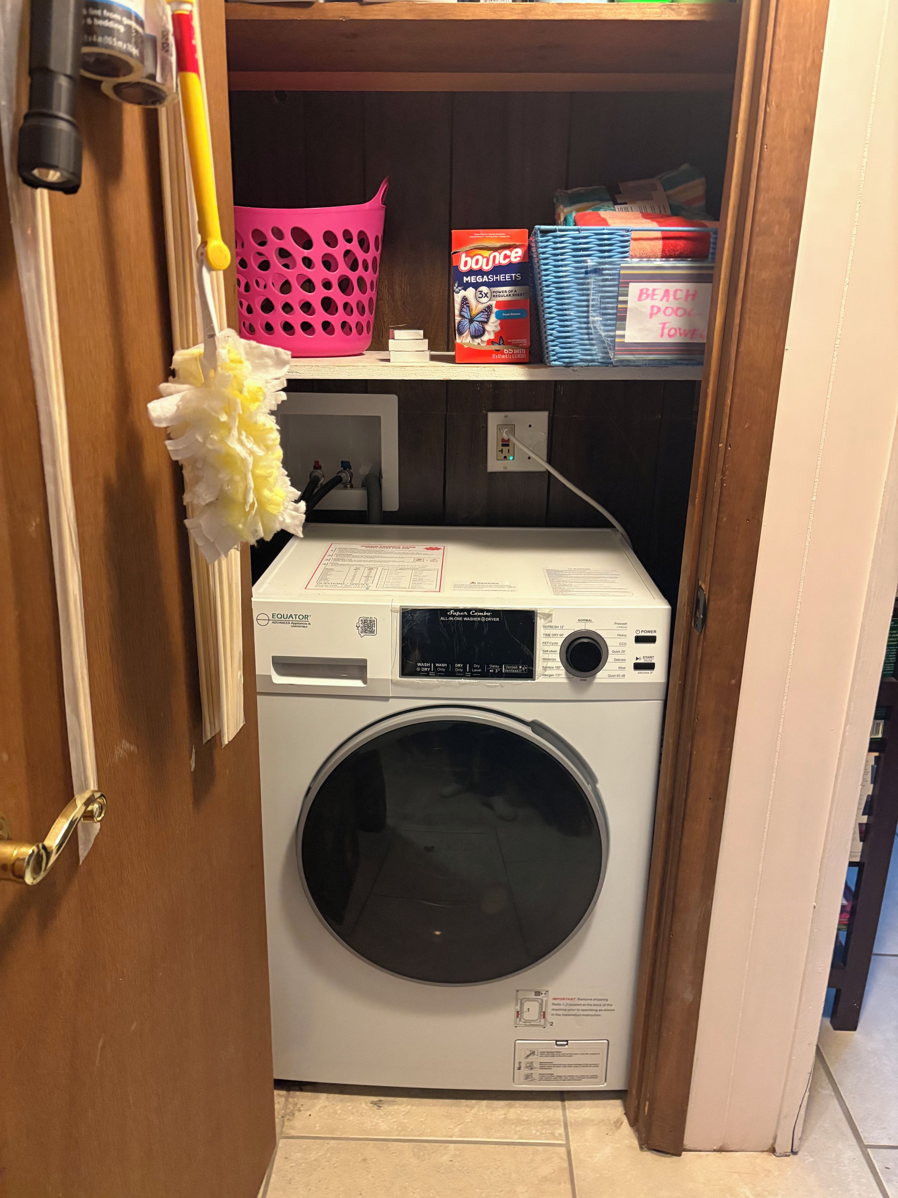 A compact, white front-loading washing machine in a laundry room with a pink basket and a box of Bounce Megasheets on the shelf.