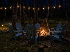 A campfire with four Adirondack chairs arranged around it in a forested area.