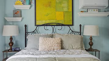 A bedroom with a bed, two bedside tables, a wall-mounted air conditioner, and a large abstract painting.