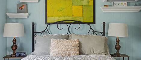 A bedroom with a bed, two bedside tables, a wall-mounted air conditioner, and a large abstract painting.