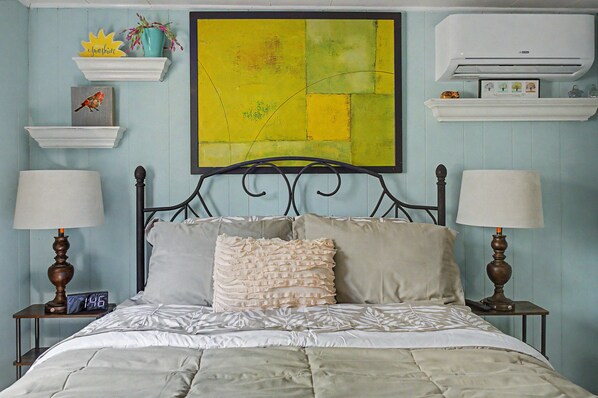 Une chambre à coucher comprenant un lit, deux tables de chevet, un climatiseur fixé au mur et un grand tableau abstrait.