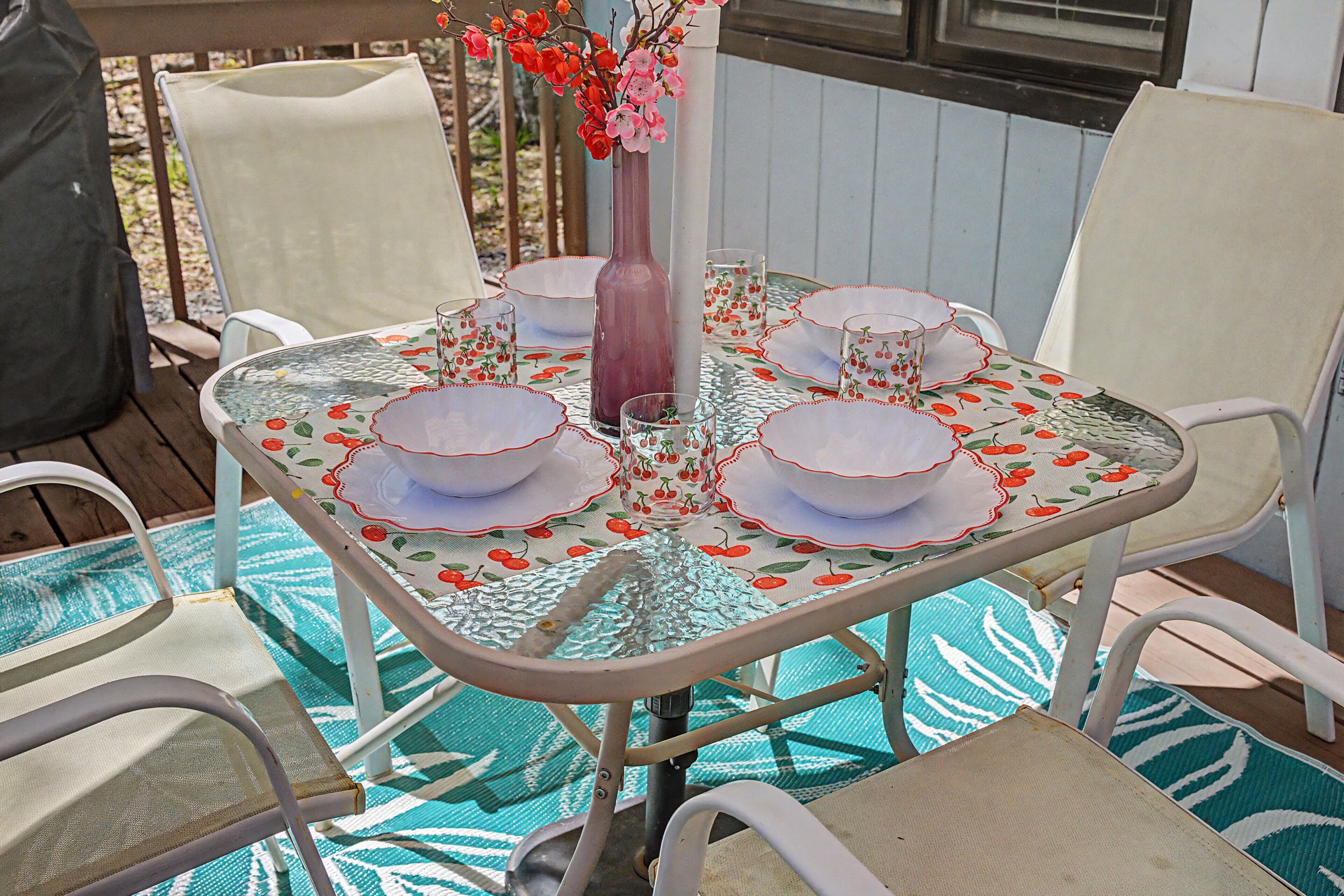 A table set with white plates, glasses, and a vase with flowers, surrounded by white chairs on a patterned tablecloth.