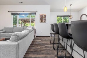 A modern living room with a grey sofa, a dining area with black chairs, and a wooden floor. - Jack’s Den | Sauna+Cold Plunge+A/C | Mt Rainier (Packwood)