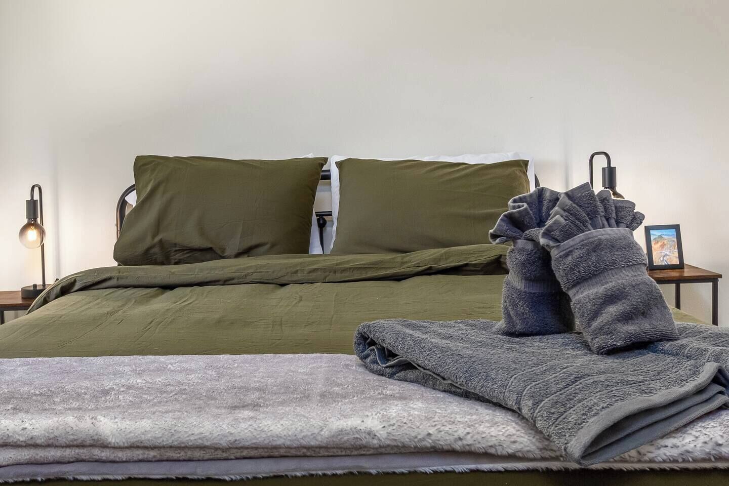 A neatly made bed with green bedding, a gray blanket, and a pair of folded gray slippers.