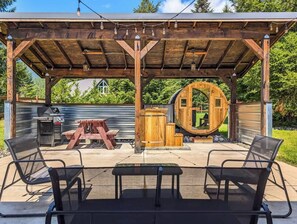 Een terras buiten met een houten pergola, een ronde houten constructie en een barbecue.