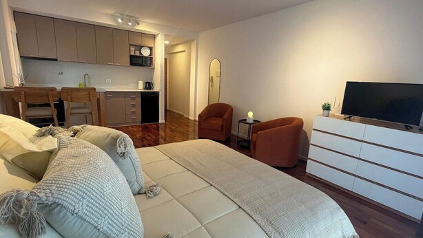 Ein modernes Schlafzimmer mit Bett, Fernseher, Kommode und einer Küche im Hintergrund.