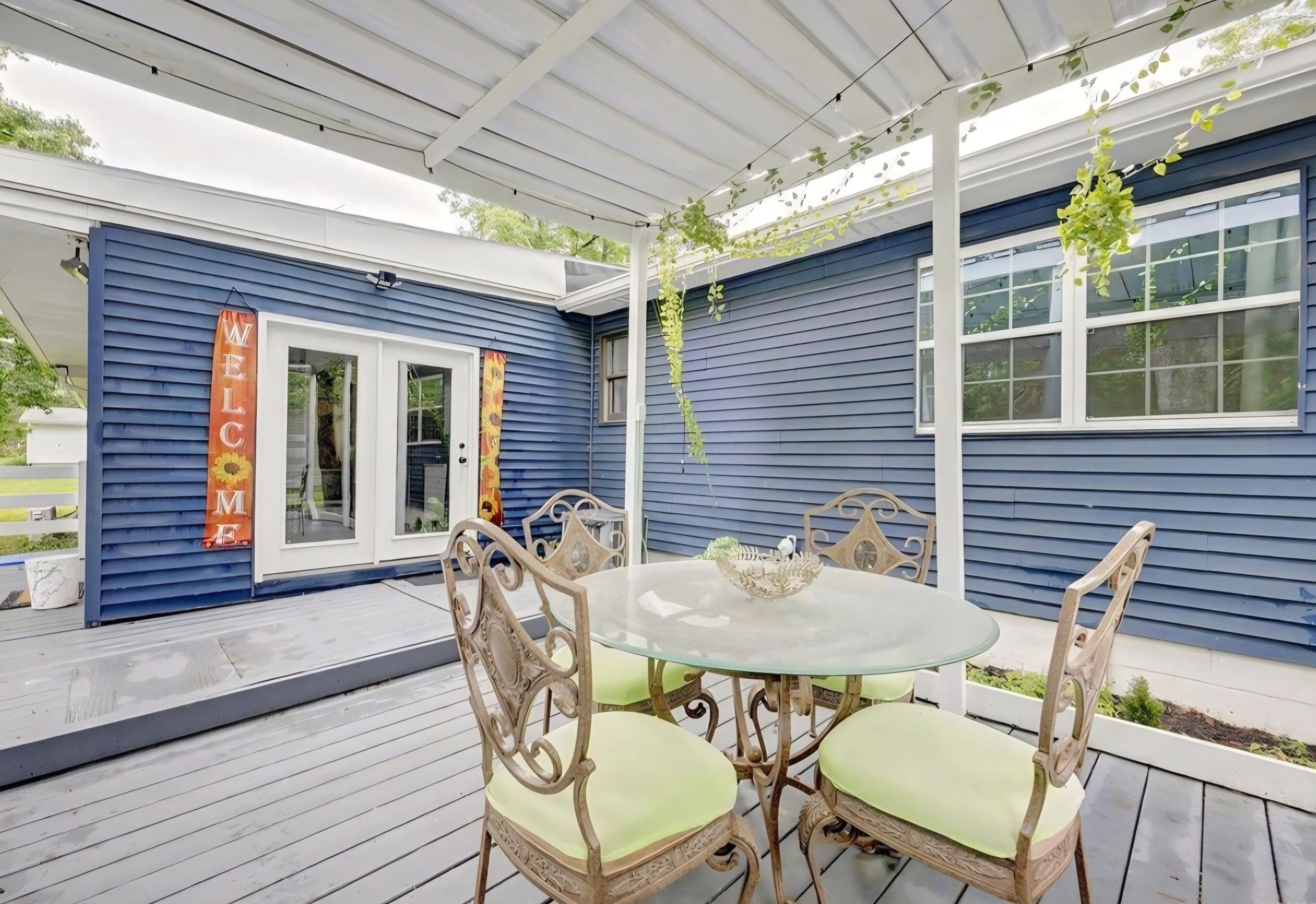 Un patio couvert avec une table ronde en verre et quatre chaises, une pergola blanche au-dessus, et un bâtiment bleu sur lequel on peut lire l’inscription « BIENVENUE ».