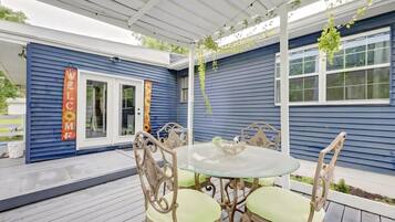 Un patio couvert avec une table ronde en verre et quatre chaises, une pergola blanche au-dessus, et un bâtiment bleu sur lequel on peut lire l’inscription « BIENVENUE ».
