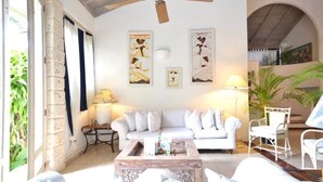 Sala de estar com sofá branco, mesa de centro e ventilador de teto. Há duas luminárias de parede e um relógio.