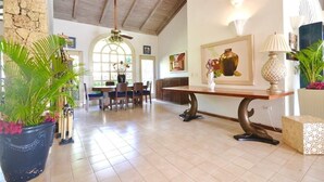 Quarto espaçoso com área de jantar, uma grande planta em vaso, mesa com arranjo floral e ventilador de teto.
