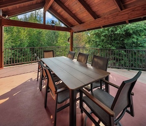 A covered outdoor dining area with a wooden table and chairs, surrounded by a metal railing, overlooking a forested area. - Private Mountain Chalet | Timberline | Mt. Rainier (Packwood)