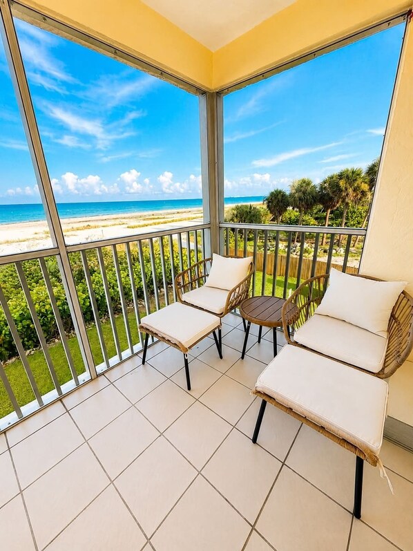 A balcony with a view of a beach, two wicker chairs with cushions, and a small round table.