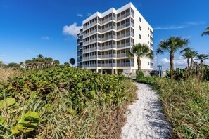 A pathway leading to a multi-story building with balconies, surrounded by dense vegetation and palm trees.