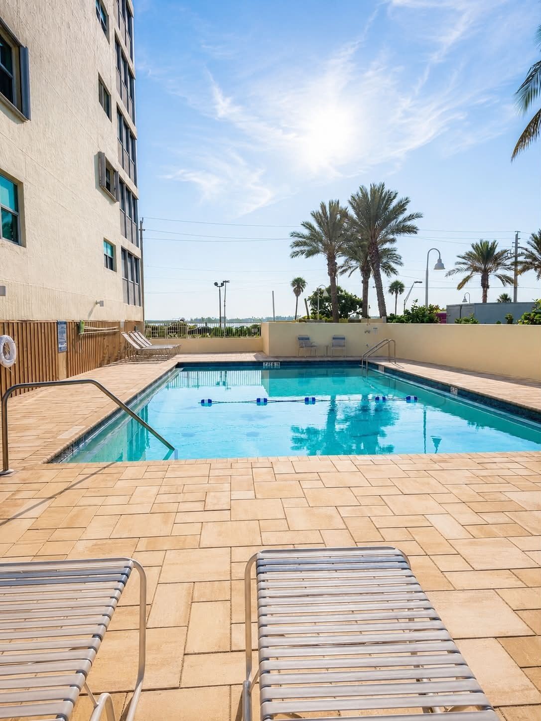 A swimming pool with lounge chairs, palm trees, and a building in the background.