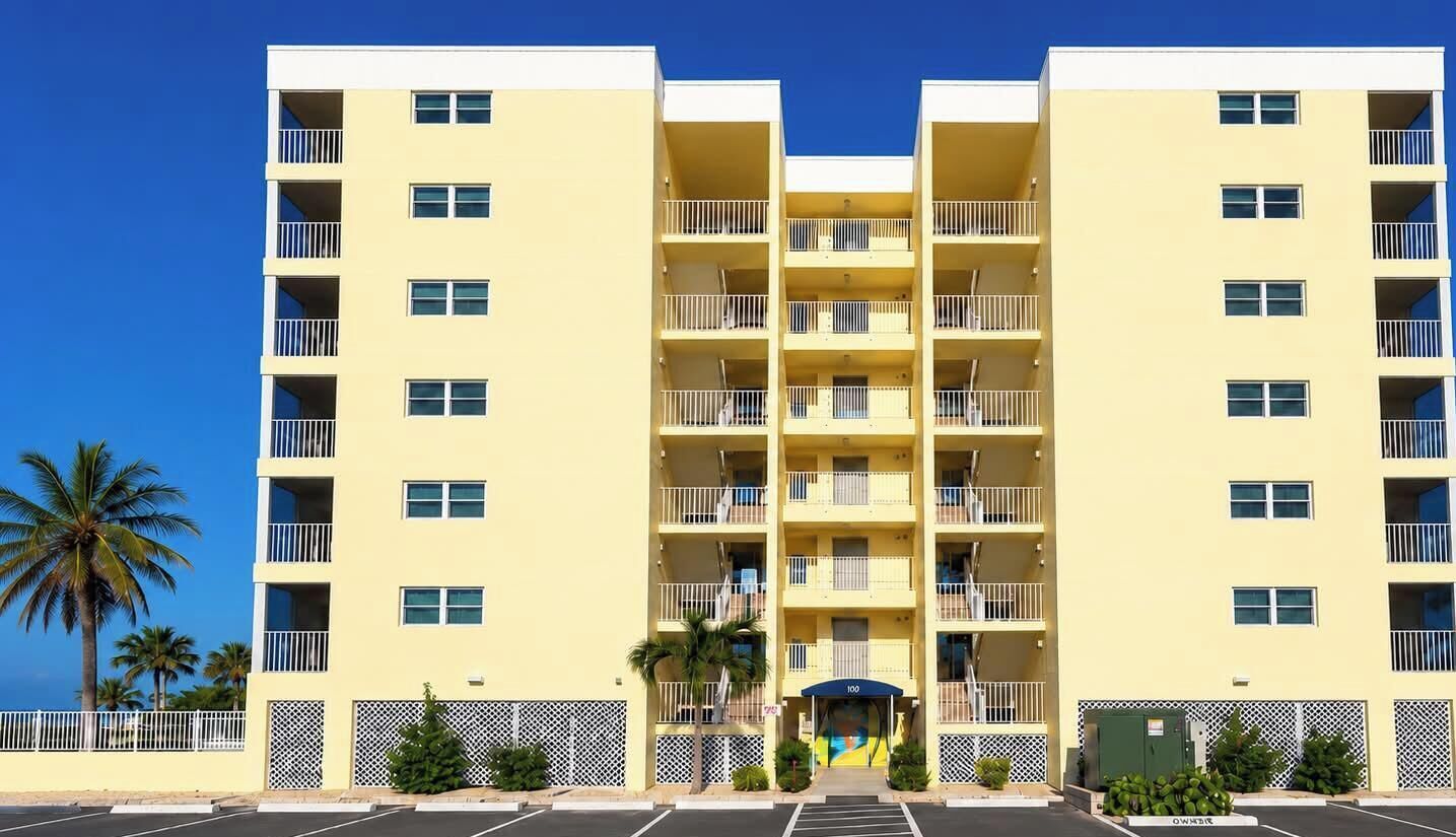 A multi-story hotel with balconies, a parking lot, and a palm tree in the foreground.