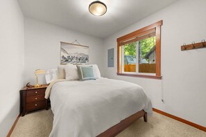 A bedroom with a large bed, a bedside table with a lamp, a window with a view of the outside, and a wall-mounted coat rack. - River Rock Retreat | Hot Tub+A/C | Mt Rainier (Packwood)