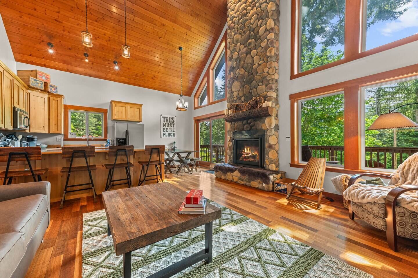 A spacious living area with a stone fireplace, wooden furniture, and large windows overlooking a forest.