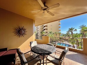 A balcony with a ceiling fan, a round metal table with chairs, a sun decoration, and a view of a swimming pool and palm trees. - Bella Sirena B-202 (Puerto Peñasco)