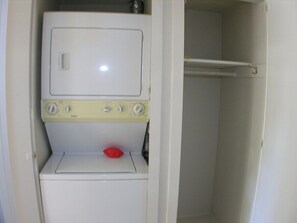 A compact, white laundry room with a washing machine and dryer, a red pillow on the dryer, and a white towel rack. - Villa De Las Flores - South Palm Springs (Palm Springs)