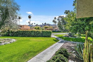 A well-maintained garden with a pathway leading to a building, surrounded by greenery and palm trees. - Villa De Las Flores - South Palm Springs (Palm Springs)