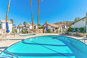 Piscina con sillas de descanso y sombrillas, rodeada de edificios de estilo mediterráneo.