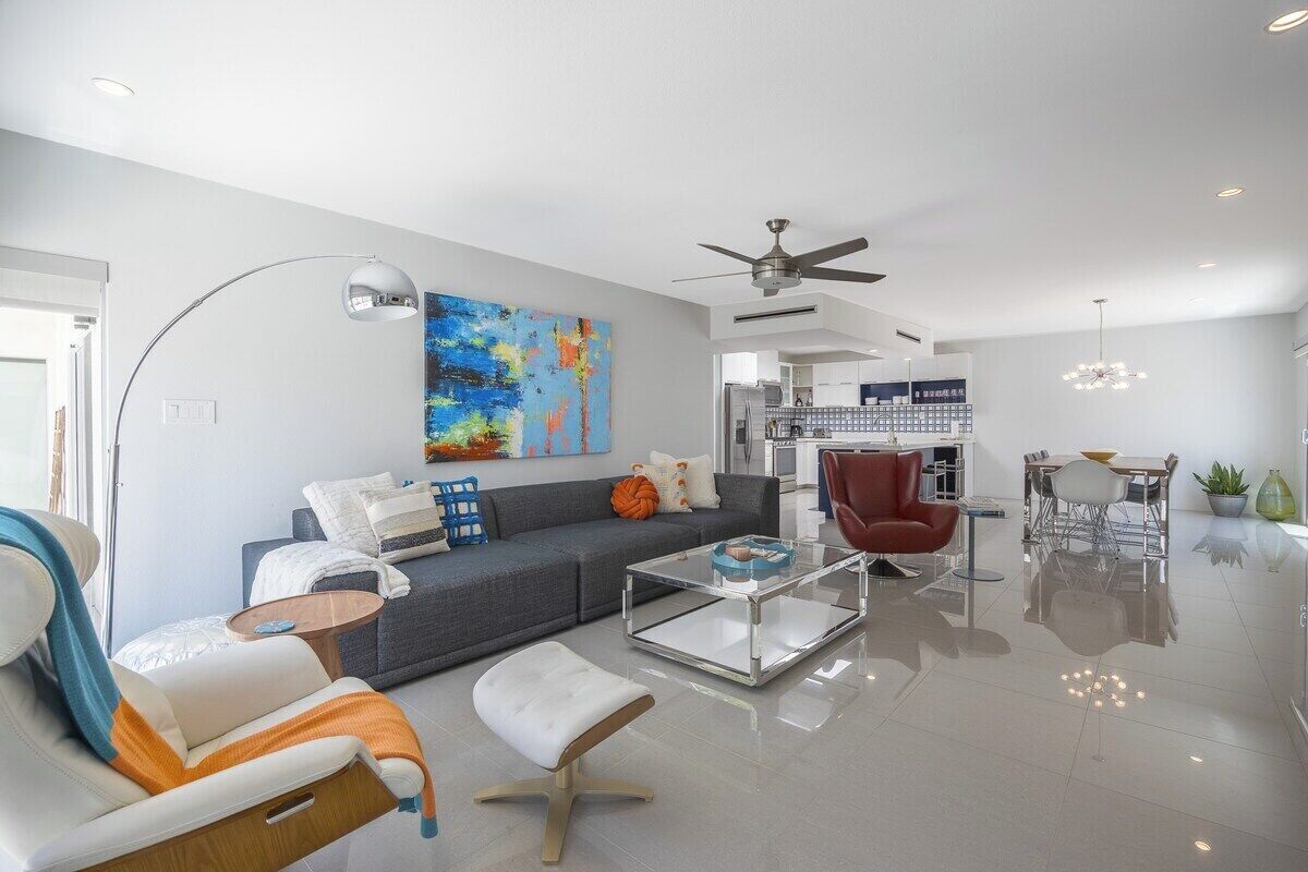 A modern living room with a sofa, armchair, and ottoman, a glass coffee table, a ceiling fan, and a dining area with chairs and a table.