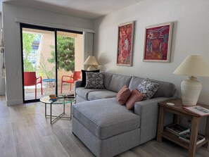 Ein Wohnzimmer mit einem grauen Sofa, einem Glastisch, einem Holztisch mit einer Lampe und zwei gerahmten Kunstwerken an der Wand.