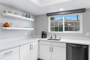 A modern kitchen with white cabinets, a coffee maker, and a window with a view of a brick wall and greenery. - Mesquite Modern Dream (Palm Springs)