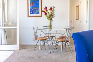 A dining area with a glass table surrounded by metal chairs with wooden seats, a vase with flowers, and a framed painting on the wall.