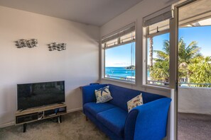 A living room with a flat-screen TV, a blue sofa, and a view of the ocean through large windows.