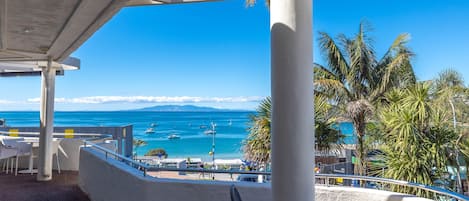 A seaside terrace with a view of the ocean and boats, featuring outdoor seating and a palm tree.