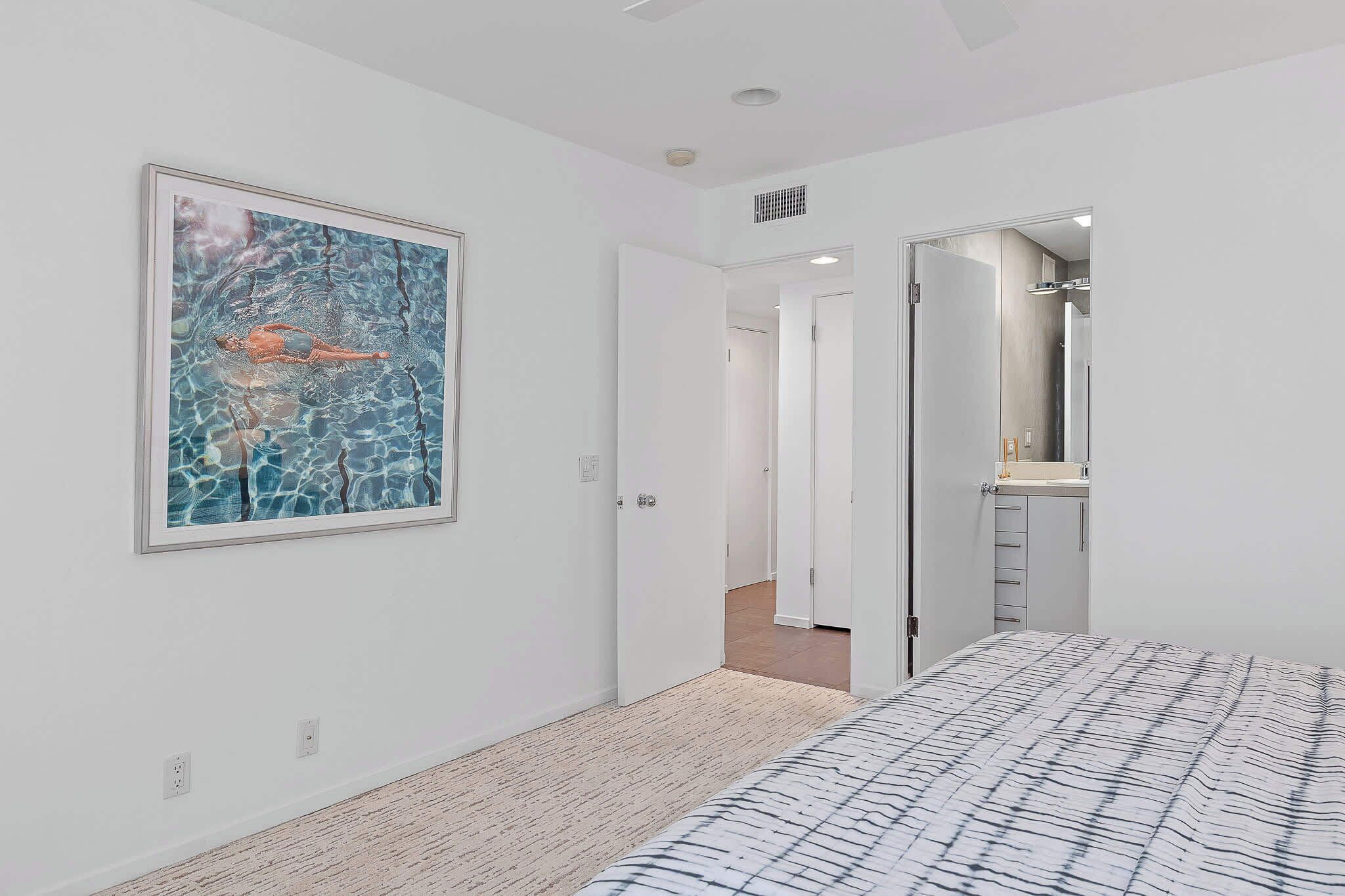 Une chambre à coucher moderne avec un grand lit, un tableau abstrait encadré et une salle de bain visible à travers une porte ouverte.