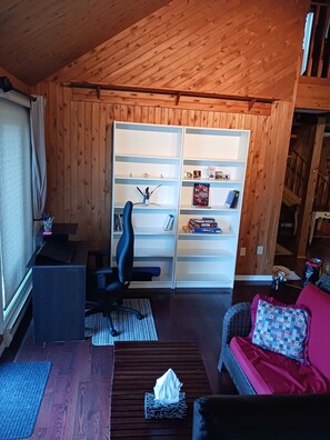 A room with a wooden ceiling, a desk with a chair, a bookshelf, a sofa with a cushion, and a patterned rug.
