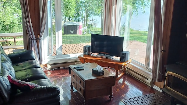Un accogliente soggiorno con divano, tavolino da caffè e televisione. Grandi finestre con tende, soffitto in legno e vista sull'esterno.