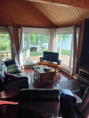 A cozy living room with a sofa, a coffee table, and a television. There are large windows with curtains, a wooden ceiling, and a view of the outdoors.