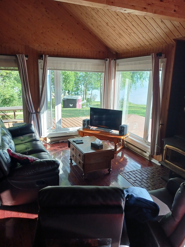 A cozy living room with a sofa, a coffee table, and a television. There are large windows with curtains, a wooden ceiling, and a view of the outdoors.