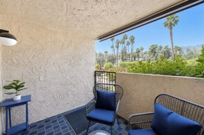 A balcony with a textured wall, two chairs with cushions, a small table with a plant, and a view of palm trees and greenery. - PS Golf and Tennis Stay (Palm Springs)