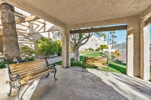 A covered outdoor seating area with benches, a concrete floor, and a view of a building and greenery. - PS Golf & Tennis Club Condo (Palm Springs)