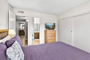 A bedroom with a bed, a dresser, a television, and a bathroom visible through an open door. - PS Golf & Tennis Club Condo (Palm Springs)
