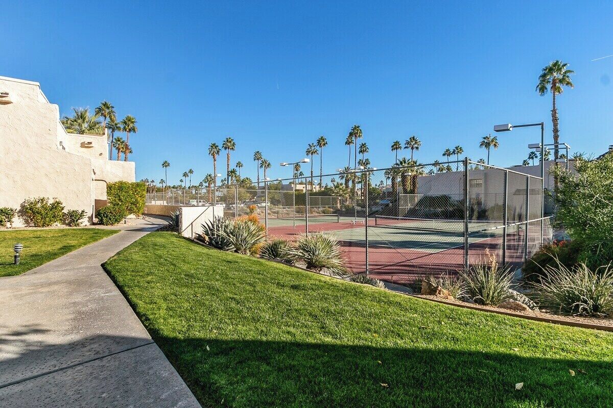 A tennis court surrounded by a fence, with a building to the left and palm trees in the background.