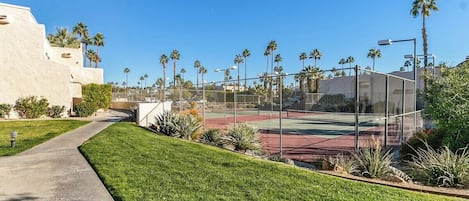 Una cancha de tenis rodeada por una valla, con un edificio a la izquierda y palmeras al fondo.
