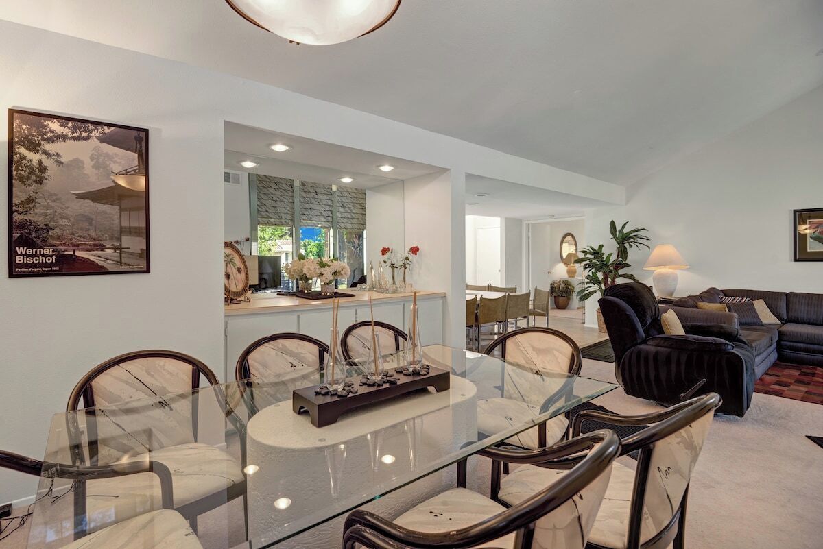 A modern dining area with a glass table and chairs, a kitchen in the background, and a living room with a sofa and lamps.