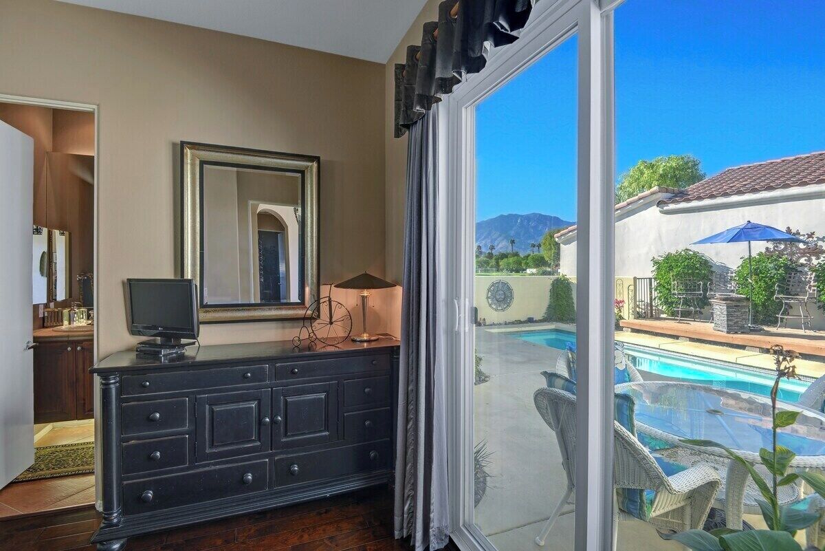A room with a large window, a dark wooden dresser, a television, and a view of a swimming pool and mountains.