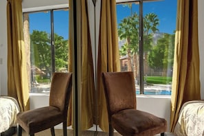 A room with two chairs facing a large window with curtains, a view of a pool and greenery outside. - Ramon Estados Treehouse (Palm Springs)