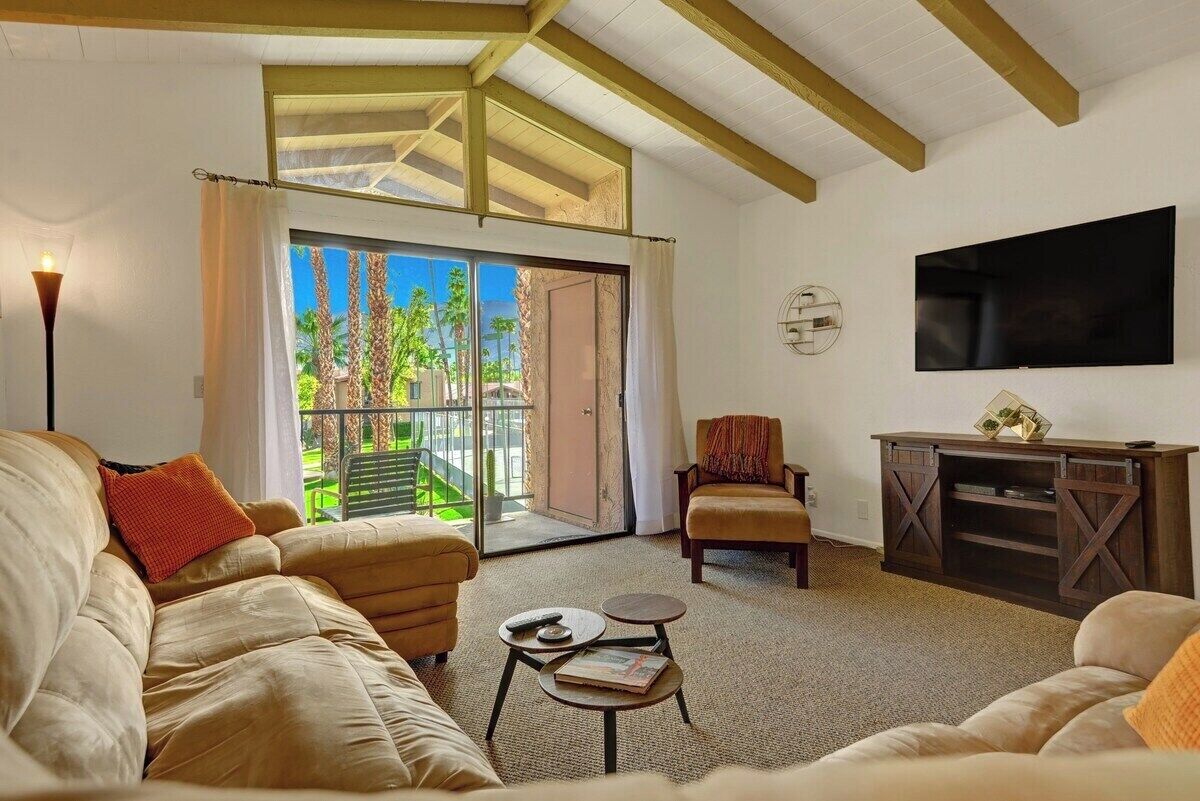 A living room with a sofa, armchair, and a TV on the wall. There is a sliding glass door leading to an outdoor area with greenery.