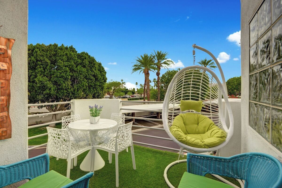 Une terrasse avec une table ronde, des chaises blanches, un fauteuil suspendu et une vue sur les arbres et les palmiers.