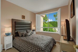 Ein Schlafzimmer mit einem großen Bett, einem Nachttisch, einer Kommode und einem Fenster mit Blick auf Grünflächen.