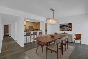 A modern dining area with a wooden table and chairs, a chandelier, and a kitchen in the background. - Garden Villas Paradise (Palm Springs)