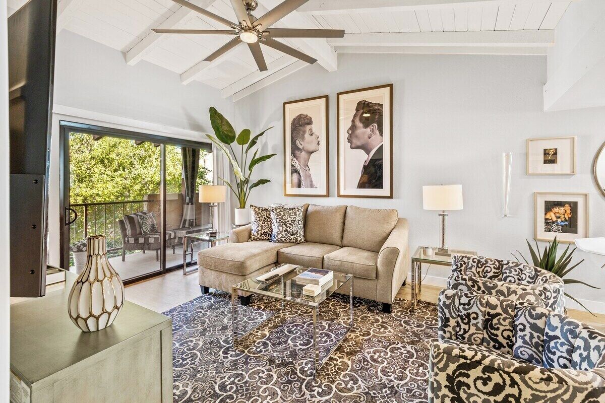 A living room with a sofa, coffee table, and patterned rug. There are two framed pictures on the wall, a ceiling fan, and a plant.